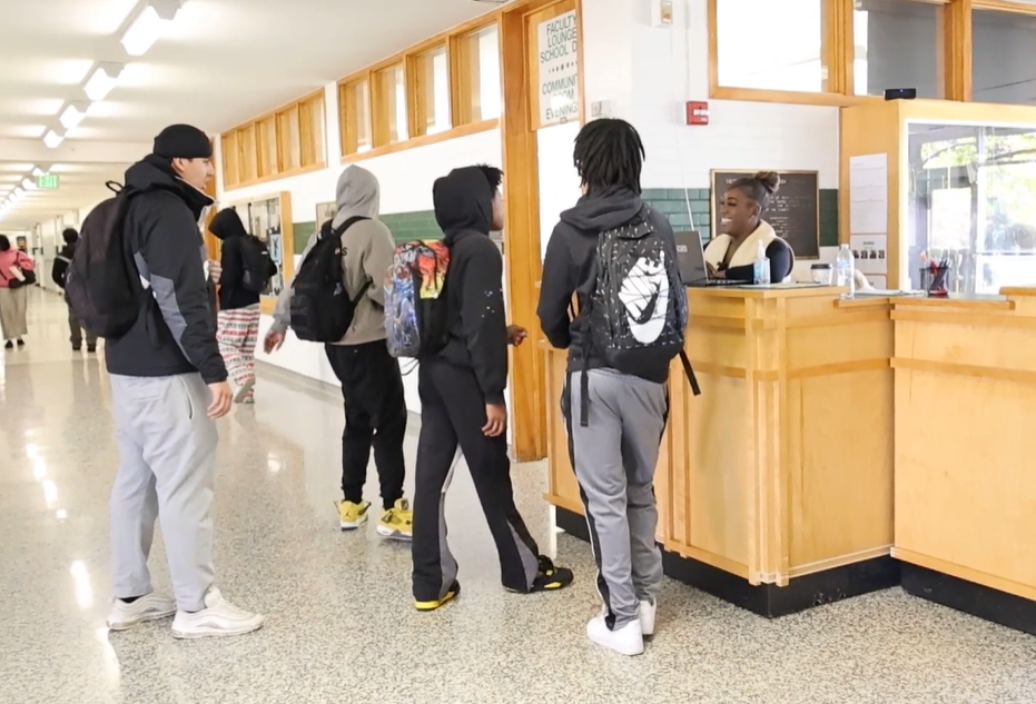 Four students speak with a woman behind a desk in a school hallway. 