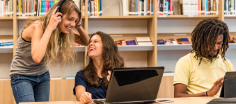 Three young, smiling adults with their laptops out having fun in the library. 