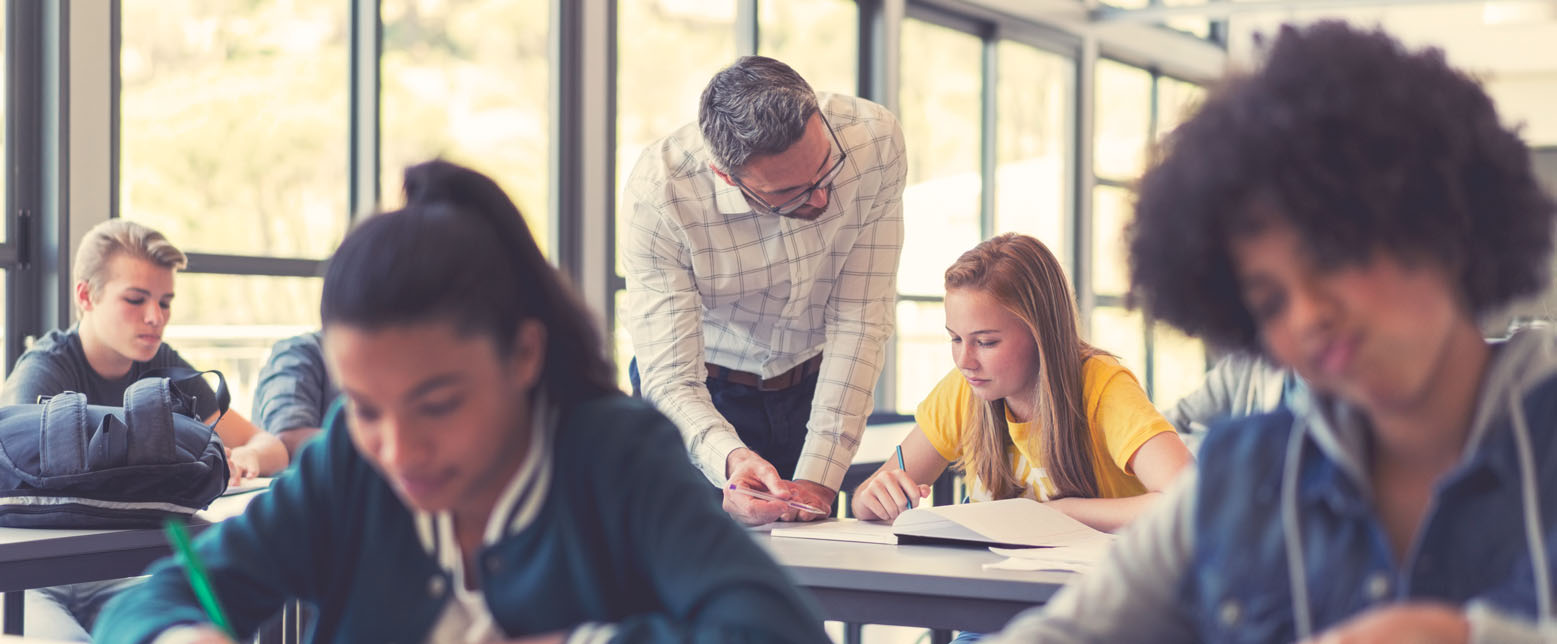 Students at desks in a classroom studying while a teacher helps